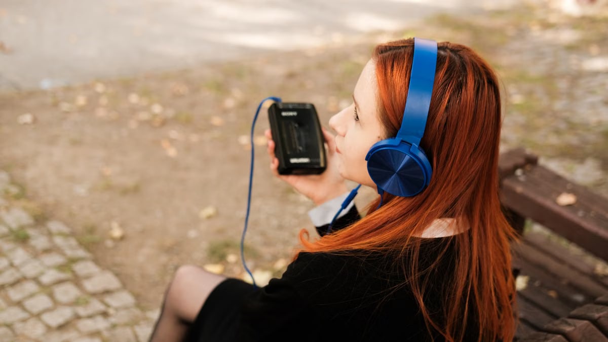 A redheaded teenager listens to music on headphones while sitting on a bench outside.